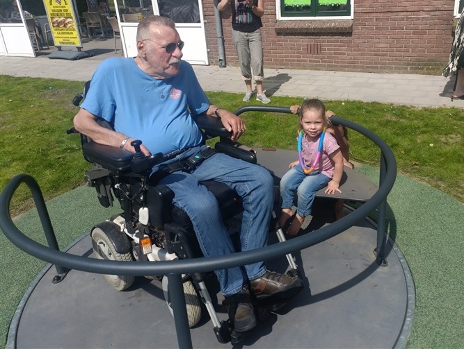 man in rolstoel samen met kleinkind op draaispeeltoestel.