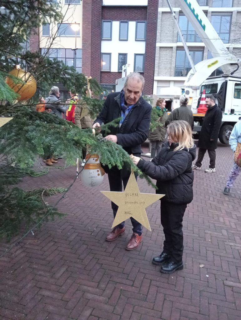 Kinderambassadeur Jasmijn en wethouder Bob Roelofs hangen een ster en sneeuwpop in de kerstboom. Op de kerstster van Jasmijn staat een wens voor ‘gelijke behandeling’ en wethouder Bob Roelofs hangt een sneeuwpop in de boom met de wens ‘dat iedereen in de speeltuin kan spelen’.